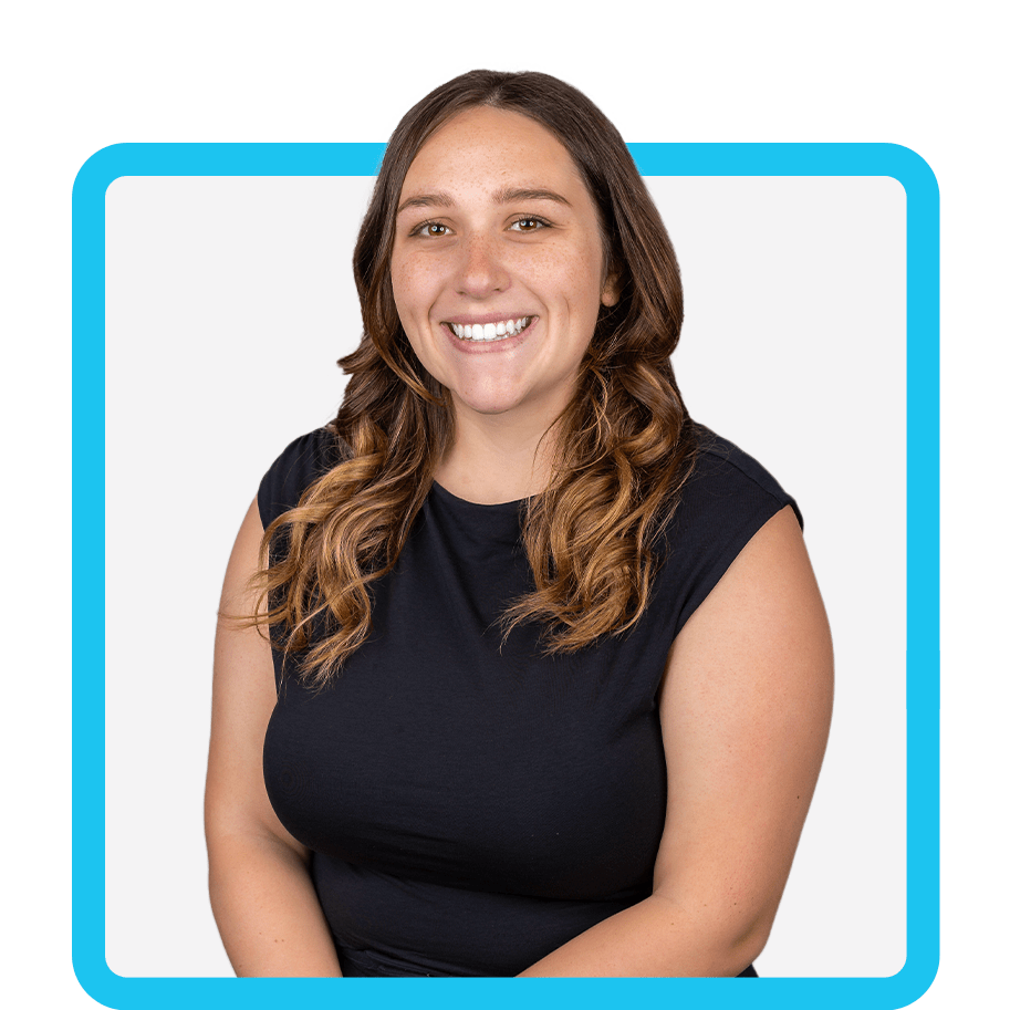 A woman with wavy brown hair wearing a black sleeveless top smiles at the camera. She is sitting in front of a plain white background, framed by a blue rectangular outline.