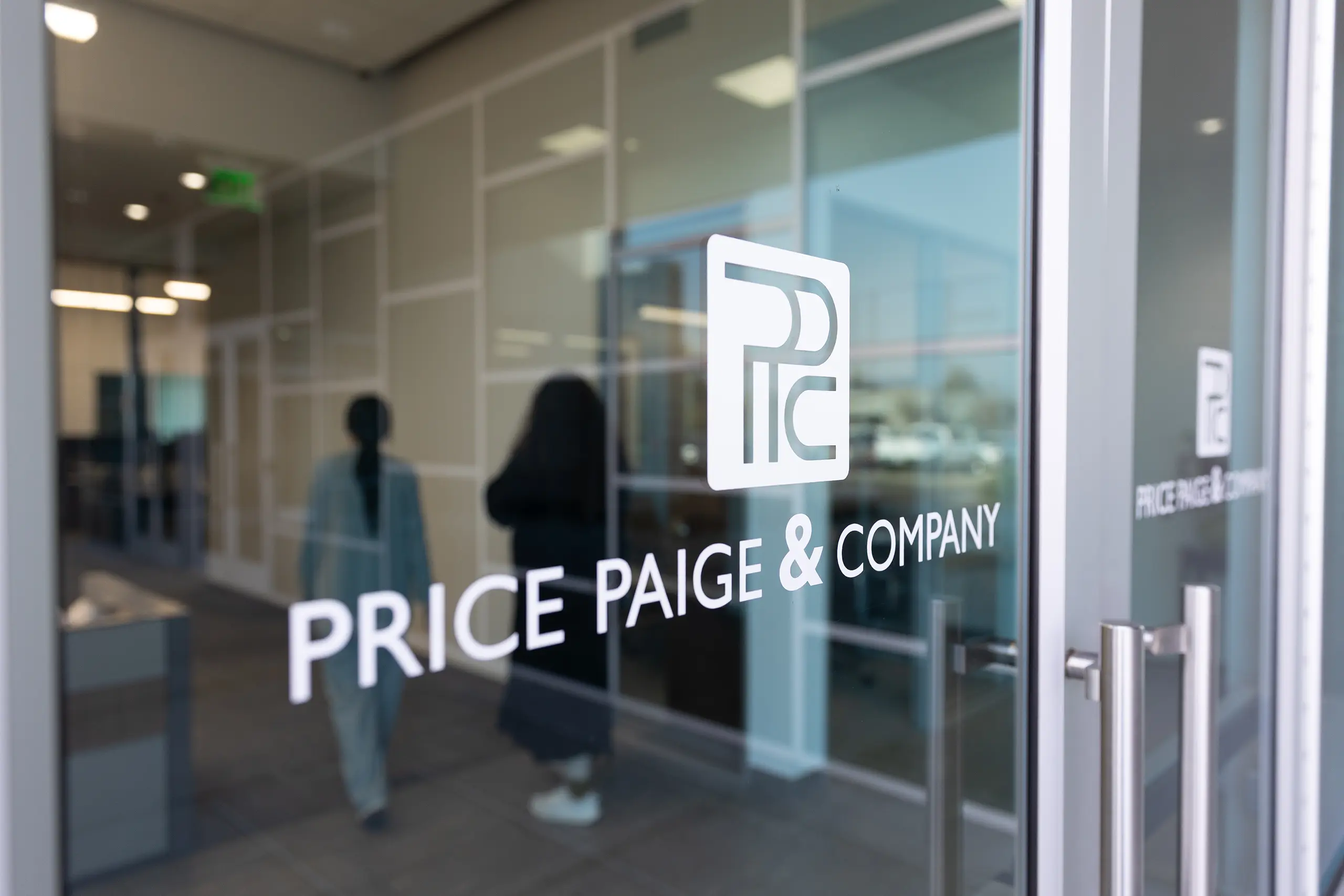 Glass door with the logo and name “PRICE PAIGE & COMPANY” in white letters; two people are walking inside an office with glass walls and natural light.